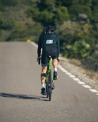 A cyclist in full gear rides along a smooth, empty road surrounded by greenery. The rider is wearing a dark uniform and helmet, showcasing an athletic posture. The bicycle has a green frame, and shadows are cast on the road surface.