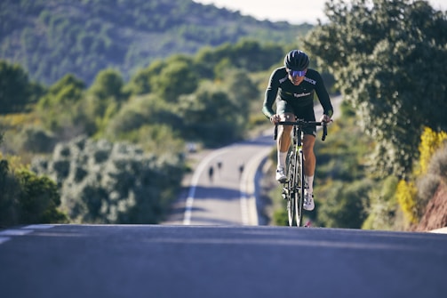 A cyclist wearing a helmet and cycling gear rides a bicycle uphill along a winding road surrounded by lush greenery and forested hills. The background features blurred scenery with another cyclist in the distance, indicating a sense of motion and speed.