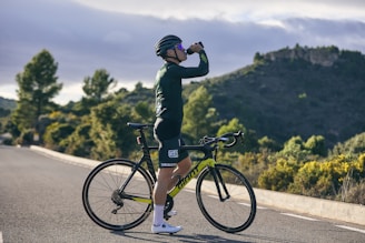A cyclist wearing a helmet and cycling gear is standing on a road, drinking from a bottle. The surrounding landscape features lush greenery and hills under a partly cloudy sky.