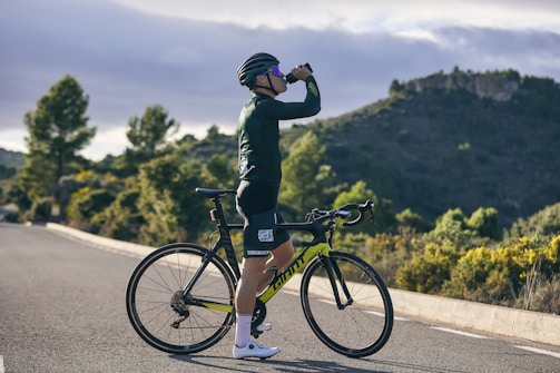 A cyclist wearing a helmet and cycling gear is standing on a road, drinking from a bottle. The surrounding landscape features lush greenery and hills under a partly cloudy sky.