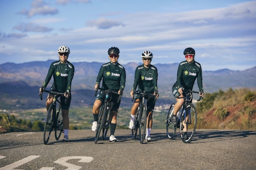 Four cyclists wearing dark green cycling jerseys and helmets are posed on road bikes, standing on a rural road with mountainous scenery in the background. The riders appear relaxed and are facing the camera, with an expansive view of nature surrounding them under a partly cloudy sky.