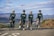 Four cyclists wearing dark green cycling jerseys and helmets are posed on road bikes, standing on a rural road with mountainous scenery in the background. The riders appear relaxed and are facing the camera, with an expansive view of nature surrounding them under a partly cloudy sky.
