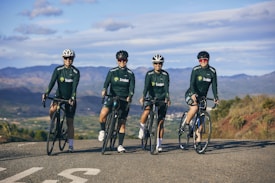 Four cyclists wearing dark green cycling jerseys and helmets are posed on road bikes, standing on a rural road with mountainous scenery in the background. The riders appear relaxed and are facing the camera, with an expansive view of nature surrounding them under a partly cloudy sky.