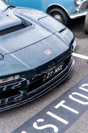 A close-up of a black Honda NSX car, adorned with raindrops and parked next to a vintage blue car. The license plate and distinct Honda logo are visible. The vehicle is parked in a space marked for visitors, with the asphalt wet from rain.