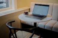 A round table with a laptop and a cup of coffee placed on it, situated in a cozy cafe setting with a cushioned bench and natural light coming through a window.