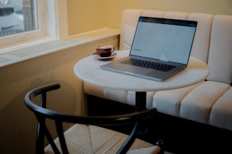 A round table with a laptop and a cup of coffee placed on it, situated in a cozy cafe setting with a cushioned bench and natural light coming through a window.