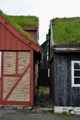 Two traditional wooden houses with grass-covered roofs are positioned closely, separated by a narrow cobblestone path. The house on the left features a combination of brick and red wooden panels, while the one on the right is painted dark grey with a white-framed window.