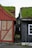 Two traditional wooden houses with grass-covered roofs are positioned closely, separated by a narrow cobblestone path. The house on the left features a combination of brick and red wooden panels, while the one on the right is painted dark grey with a white-framed window.