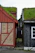 Two traditional wooden houses with grass-covered roofs are positioned closely, separated by a narrow cobblestone path. The house on the left features a combination of brick and red wooden panels, while the one on the right is painted dark grey with a white-framed window.