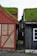 Two traditional wooden houses with grass-covered roofs are positioned closely, separated by a narrow cobblestone path. The house on the left features a combination of brick and red wooden panels, while the one on the right is painted dark grey with a white-framed window.