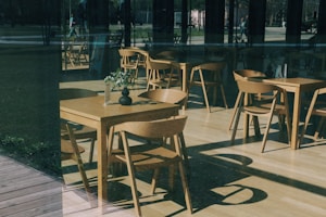 A modern caf&eacute; setting featuring wooden tables and chairs is arranged neatly inside a glass-walled space. Sunlight filters in, creating interesting shadows on the light wooden floor. A small vase with green foliage is placed on one of the tables, adding a touch of nature to the minimalist interior. Reflections on the glass display hints of the outside environment, with people and greenery faintly visible.