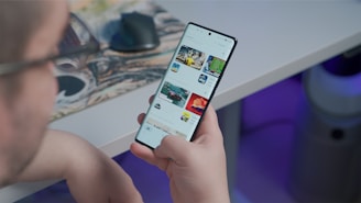 A person is holding and browsing a smartphone with a screen displaying a digital storefront or app interface. The background features a desk with a computer mouse and blurred elements, creating a tech-focused environment.
