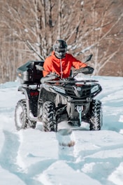 A person wearing a red jacket and a helmet is riding an all-terrain vehicle (ATV) through a snowy landscape. The ATV is black and has four large wheels. The background consists of leafless trees, suggesting a winter setting.