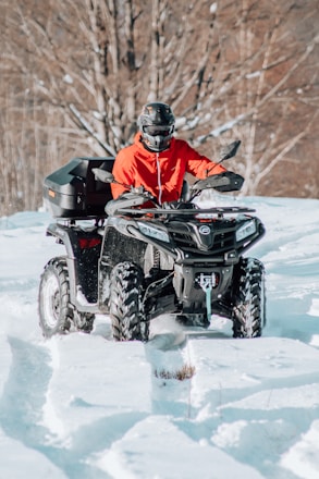 A person wearing a red jacket and a helmet is riding an all-terrain vehicle (ATV) through a snowy landscape. The ATV is black and has four large wheels. The background consists of leafless trees, suggesting a winter setting.