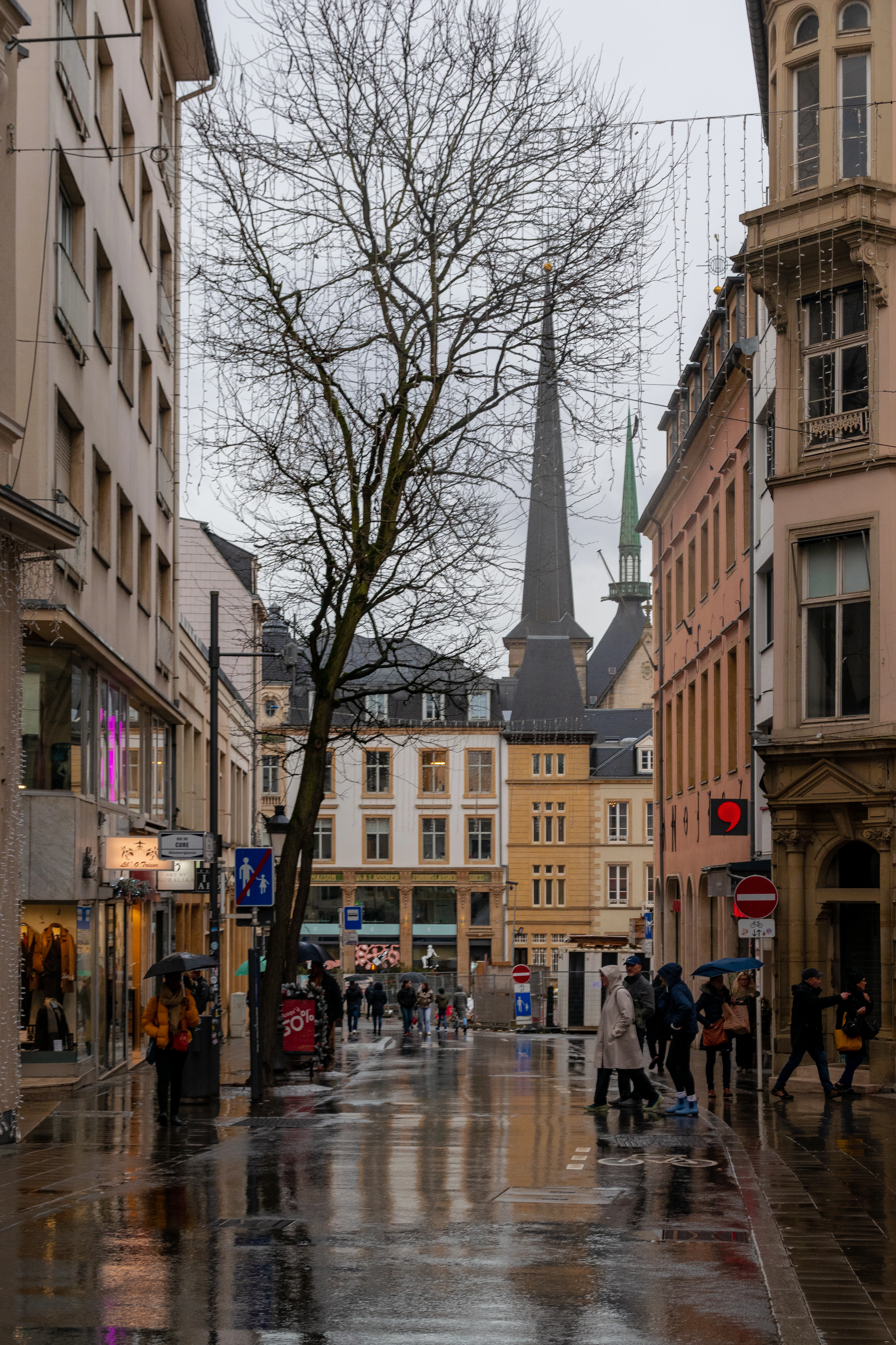 A rainy shopping street in Luxembourg City with the dark spire of Notre-Dame Cathedral rising behind bare winter trees