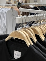 A row of wooden clothes hangers displaying various sizes of black shirts is prominently featured. In the background, more clothing items are visible, hung on a rack, showcasing a variety of colors including white and brown. The setting appears to be a retail clothing store with bright lighting.