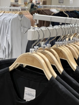 A row of wooden clothes hangers displaying various sizes of black shirts is prominently featured. In the background, more clothing items are visible, hung on a rack, showcasing a variety of colors including white and brown. The setting appears to be a retail clothing store with bright lighting.