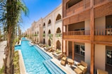 A luxurious building with an ornate design featuring arches and Arabic-inspired architecture is complemented by a long, narrow swimming pool running parallel to it. The pool is surrounded by palm trees and outdoor seating areas with wicker furniture, offering a relaxing ambiance. The sky is clear and vividly blue, reflecting off the pool's water.