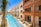 A luxurious building with an ornate design featuring arches and Arabic-inspired architecture is complemented by a long, narrow swimming pool running parallel to it. The pool is surrounded by palm trees and outdoor seating areas with wicker furniture, offering a relaxing ambiance. The sky is clear and vividly blue, reflecting off the pool's water.