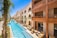 A luxurious building with an ornate design featuring arches and Arabic-inspired architecture is complemented by a long, narrow swimming pool running parallel to it. The pool is surrounded by palm trees and outdoor seating areas with wicker furniture, offering a relaxing ambiance. The sky is clear and vividly blue, reflecting off the pool's water.