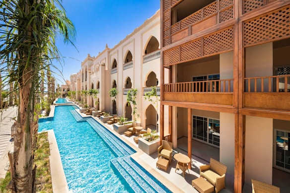 A luxurious building with an ornate design featuring arches and Arabic-inspired architecture is complemented by a long, narrow swimming pool running parallel to it. The pool is surrounded by palm trees and outdoor seating areas with wicker furniture, offering a relaxing ambiance. The sky is clear and vividly blue, reflecting off the pool's water.