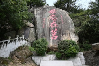 Large rock formation with pink Chinese characters etched into the surface. The rock is surrounded by lush green foliage and a stone pathway with railing leading upwards on the left.