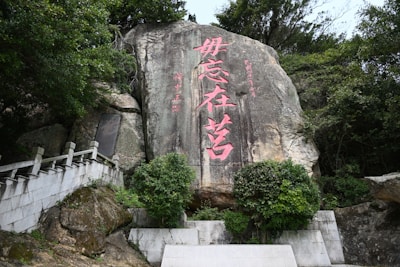 Large rock formation with pink Chinese characters etched into the surface. The rock is surrounded by lush green foliage and a stone pathway with railing leading upwards on the left.