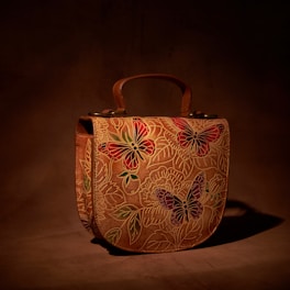 A leather handbag with intricate butterfly and floral designs, predominantly in shades of red, green, and purple. The background is a rich brown, giving a natural, earthy feel to the piece.