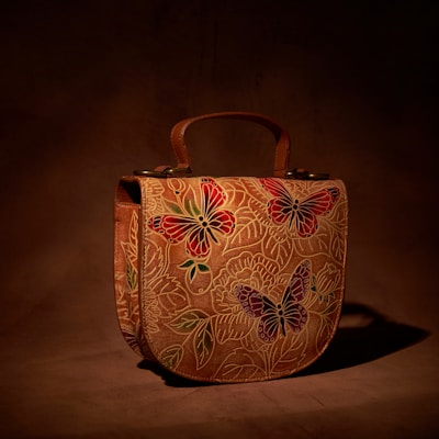 A leather handbag with intricate butterfly and floral designs, predominantly in shades of red, green, and purple. The background is a rich brown, giving a natural, earthy feel to the piece.