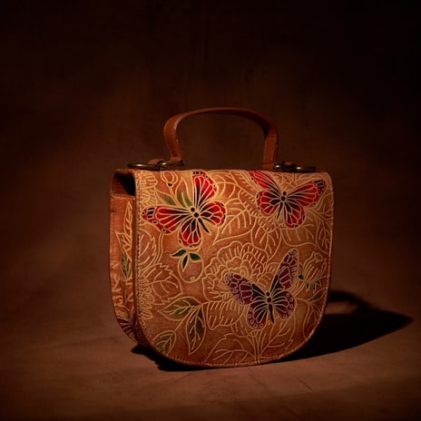 A leather handbag with intricate butterfly and floral designs, predominantly in shades of red, green, and purple. The background is a rich brown, giving a natural, earthy feel to the piece.