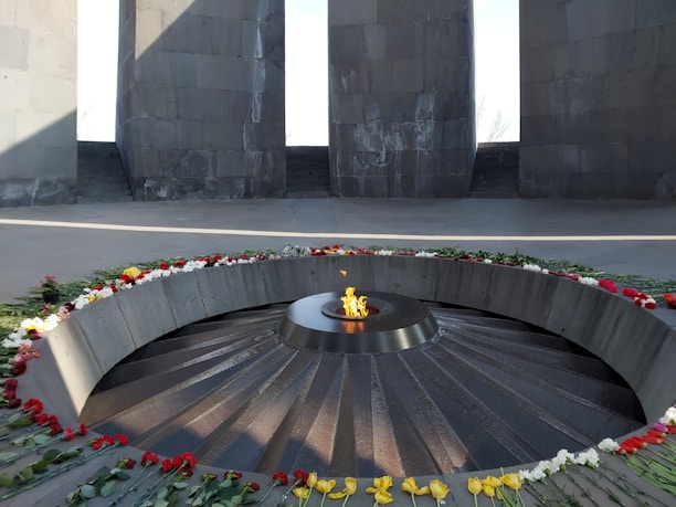 A circular eternal flame burns at the center of a memorial surrounded by a stone structure with tall columns. The flame is encircled by a bed of flowers, including roses and other colorful blooms. The setting appears somber and reverent, with natural light illuminating part of the scene and casting shadows.