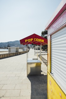 A deserted popcorn stand with a red canopy and yellow lettering is positioned on a wide, paved walkway. The stand is metallic and placed beside a yellow structure, possibly a food truck. The scene is sunny, with clear blue skies, and there are mountains and sparse trees visible in the background.