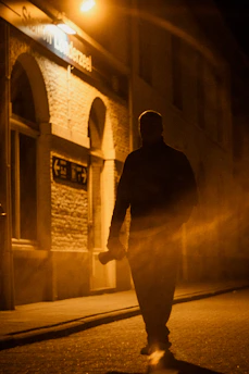 A person walking confidently at night with a focused expression, streetlights casting shadows.