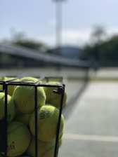 A metal basket filled with yellow tennis balls is positioned on an outdoor tennis court, with a blurred background of the net and greenery.