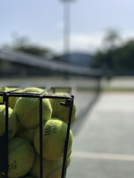 A metal basket filled with yellow tennis balls is positioned on an outdoor tennis court, with a blurred background of the net and greenery.