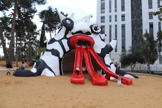 A whimsical playground structure resembling a fantastical creature with black and white patterns. It features bright red tongue-like slides. Several children are climbing and sliding on it. The surrounding area includes trees and a tall building in the background.