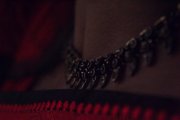 A close-up of a dark metallic necklace lying against a skin tone, with a fabric featuring red and black stripes in the background. The necklace has intricate, small details that resemble traditional textures.