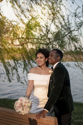 A couple in wedding attire is standing beside a wooden bench, smiling and looking into the distance. The woman is holding a bouquet of flowers. The background features a body of water and lush greenery from overhanging branches, suggesting a serene outdoor setting.