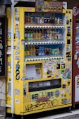 A yellow vending machine covered with various colorful stickers and graffiti, offering a variety of drinks displayed across multiple shelves. The drinks include different types and brands, and there are numerous Japanese labels and logos on the machine.