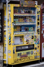 A yellow vending machine covered with various colorful stickers and graffiti, offering a variety of drinks displayed across multiple shelves. The drinks include different types and brands, and there are numerous Japanese labels and logos on the machine.