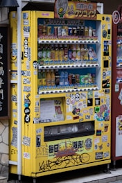 A yellow vending machine covered with various colorful stickers and graffiti, offering a variety of drinks displayed across multiple shelves. The drinks include different types and brands, and there are numerous Japanese labels and logos on the machine.