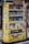 A yellow vending machine covered with various colorful stickers and graffiti, offering a variety of drinks displayed across multiple shelves. The drinks include different types and brands, and there are numerous Japanese labels and logos on the machine.