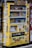 A yellow vending machine covered with various colorful stickers and graffiti, offering a variety of drinks displayed across multiple shelves. The drinks include different types and brands, and there are numerous Japanese labels and logos on the machine.