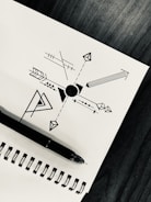A geometric design is drawn on a white sheet of paper, featuring a central circle with various arrows, lines, and shapes extending outward. A black ink pen rests on the spiral binding of the notebook, which is placed on a dark wooden surface.
