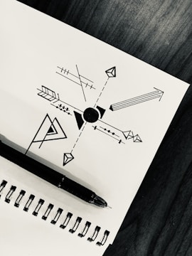 A geometric design is drawn on a white sheet of paper, featuring a central circle with various arrows, lines, and shapes extending outward. A black ink pen rests on the spiral binding of the notebook, which is placed on a dark wooden surface.