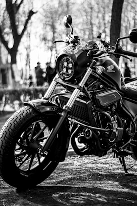 A close-up of a sleek motorcycle captured in black and white, showing its detailed components such as the front wheel and handlebars. The image features blurred trees in the background, suggesting an outdoor setting.