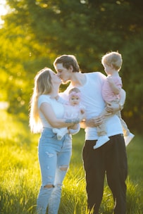 A family is standing in a sunlit field. The adults are sharing a kiss while holding a toddler and a baby. The sunlight creates a warm glow around them, highlighting their expressions. The father's head is turned towards the mother, and they are surrounded by green foliage.