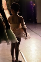 A young ballet dancer stands in the wings of a stage wearing a peach-colored tutu with lace detailing. Her hair is neatly tied up, and she is captured in a moment of anticipation. The stage lights create a warm glow around her and highlight the delicate fabric of her costume. Another dancer is partially visible, dressed in a pale green tutu.