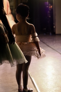 A young ballet dancer stands in the wings of a stage wearing a peach-colored tutu with lace detailing. Her hair is neatly tied up, and she is captured in a moment of anticipation. The stage lights create a warm glow around her and highlight the delicate fabric of her costume. Another dancer is partially visible, dressed in a pale green tutu.
