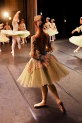 Several ballet dancers on stage wearing elegant, classical costumes are captured in motion under warm stage lighting. The dancers are dressed in pastel-colored tutus and are engaged in a graceful performance. One dancer in the foreground is standing in a poised position, while others in the background are captured mid-spin.